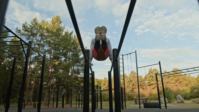 Low angle shot of young man in sports wear performing L-sit on parallel dip bars in outdoor training area in forest