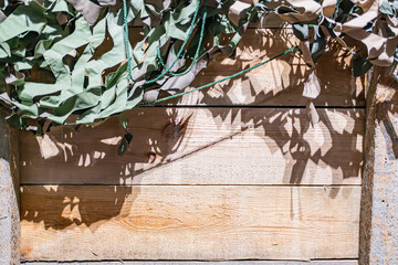 Military camouflage net, rustic wooden structure with natural covering, outdoor setting, sunlight streaming, dappled shadows, daytime, upward view, wood planks, vegetation or vines, candid capture, e