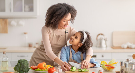 Domestic Life. Portrait of smiling African American woman helping daughter to prepare fresh healthy vegetable salad, little girl learning how to cook, chopping cucumber with knife on cutting board