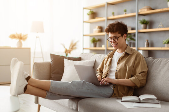 Young casual asian guy using laptop sitting on sofa in living room, free copy space. Happy man in eyeglasses chatting with friends online, male freelancer working from home, typing on keyboard