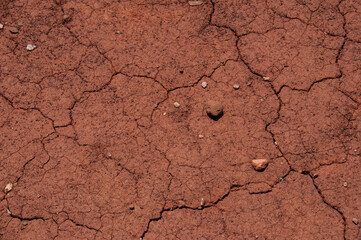A detailed, top-down close-up of dry, cracked red earth with a network of fine lines and a few small rocks scattered across the surface.