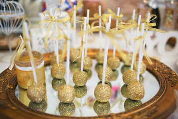 Golden glitter cake pops with ribbons on sticks, displayed on a mirror tray. Elegant and festive desserts for weddings and glamorous events.