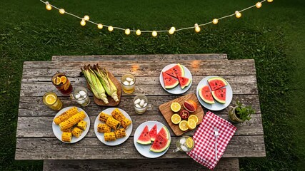 Rustic outdoor picnic table spread with watermelon, corn, and string lights