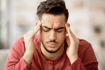Closeup of bearded arab guy suffering from migraine at home, touching his temples with closed eyes,...