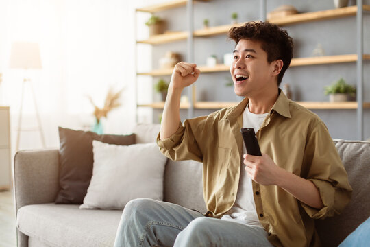 Portrait of excited young guy watching football match, shaking clenched fist. Happy asian man relaxing sitting on couch cheering favorite team enjoying game on TV at home holding remote controller - Powered by Adobe