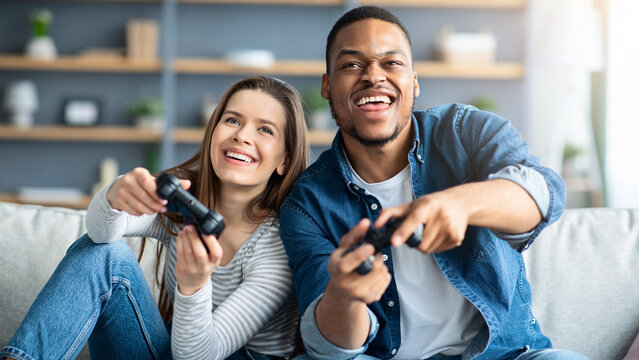 Domestic Entertainment. Joyful young multicultural couple playing video games in living room, cheerful interracial lovers holding joysticks and having fun together at home, competing with each other