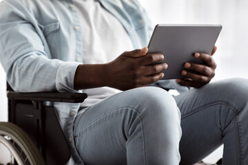 Surfing in internet, watch video, make online calls and work remotely during COVID. Adult african american disabled guy in wheelchair, uses tablet for chat in living room or clinic interior, cropped