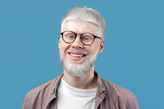 Headshot portrait of happy albino blond man with pale skin wearing eyeglasses, looking and smiling at camera against turquoise studio background. Excited guy with unusual appearance posing in specs