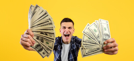 Excited hipster guy holding fans of money, winning lottery or casino bet on orange studio background. Funky millennial man with dollars reaching financial success, cannot believe his luck
