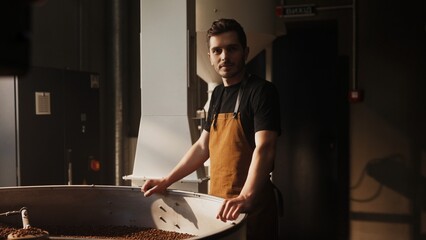 Caucasian male with short brown hair wearing black shirt and brown apron. Standing near cooling tray with roasted coffee beans. Looking directly at camera. Overseeing roasting process with confidence.