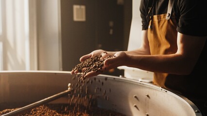 Bearded Caucasian man wearing brown apron holding roasted coffee beans in both hands. Observing texture and aroma while checking roast quality during coffee production inside workshop.
