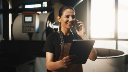 Focused Caucasian woman in brown apron holding tablet while talking on smartphone. Standing near roasting equipment and checking production details in bright coffee processing workspace.