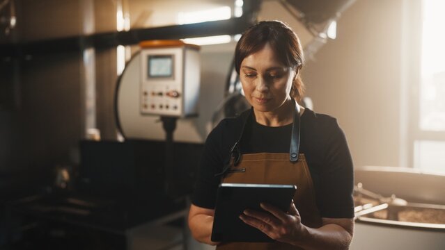 Focused beautiful Caucasian woman with tied dark hair wearing brown apron. Holding tablet in hands. Standing near coffee roasting equipment and monitoring production process in bright factory room.