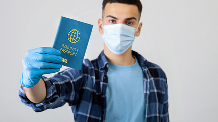Young guy in medical mask showing covid-19 immunity passport on light studio background, selective focus. Millennial man with vaccination certificate to prevent coronavirus spread