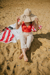 A woman is sitting on the beach wearing a straw hat and red top