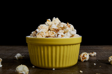 Yellow bowl with popcorn on wood, deep black background. Centered composition, with less dispersion. Ideal for product photography or minimalist food content.