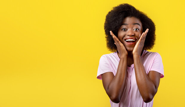 Breathtaking advertisement. Emotional african american lady with hands on cheeks looking at camera over yellow background, studio shot. Wow offer concept