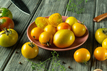 Varieties of yellow tomatoes are gathered on a pink plate with a backdrop of fresh herbs and a knife resting nearby, creating a vibrant and inviting kitchen atmosphere