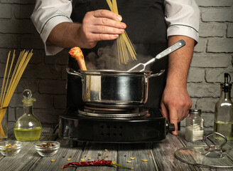 A chef prepares to cook pasta by adding dry spaghetti to boiling water in a metal pot. Surrounding ingredients include olive oil, spices, and herbs on a rustic table