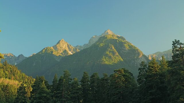 Majestic mountains rise against a clear blue sky, adorned with patches of greenery. Tall trees frame the base, creating a serene and picturesque natural landscape