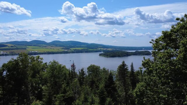Slow flight through the treetops of a forest toward an island in the center of a large lake in a rural agricultural setting under a cloudy sky. La Matapedia, Quebec, Canada, 2025.