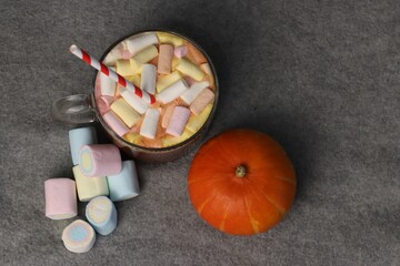 Cozy autumn photo: glass cup of hot cocoa with marshmallows and festive striped straw next to bright pumpkin — great choice for advertising layouts, coffee shop menus, fall holiday greeting cards