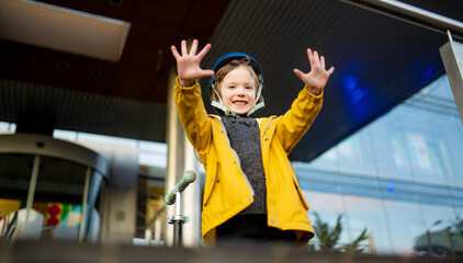 Cheerful child raises both hands in excitement while standing by their scooter, dressed in a yellow...