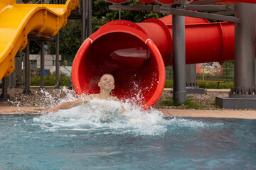 Joyful child at bottom of red tube slide with eyes closed and arms wide open enjoying huge water...
