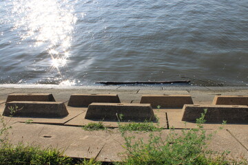 Calm Waters Reflecting Sunlight Along a Serene Riverbank With Patterned Concrete Blocks and Lush Greenery in a Peaceful Outdoor Setting