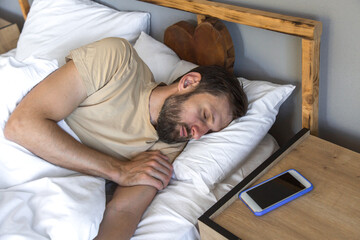 Man sleeping peacefully in bed next to smartphone on wooden nightstand, concept of digital detox and sleep quality.