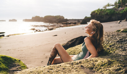 Woman reclines with earbuds on mossy rock, face turned to sun, merging natural grounding with digital audio escape, expressing balance between technology and personal tranquility.