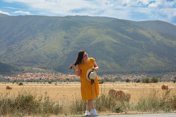 Girl in yellow dress with hat in hands in glasses against mountains in Bulgaria. Beautiful landscape