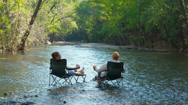Friends enjoying relaxing moments in river. Friends relaxing on camping chairs midstream, sipping drinks and chatting while surrounded by lush riverside landscape during peaceful summer getaway moment