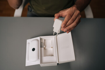 High-angle view of unrecognizable man unpacking box, holding white smartphone charger being unpacked from cardboard box, sitting on gray table. Concept of contemporary gadget unboxing.
