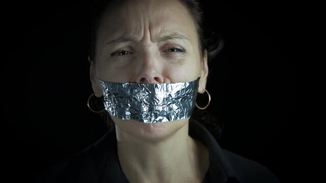 Female hostage with closed mouth. A view of sad woman hostage with take on her mouth to stop speaking against black background