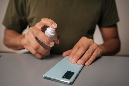Sanitizing smartphone surface, male hands applying disinfectant spray for preventing viral transmission and maintaining personal hygiene sitting at table. Concept of healthy lifestyle.
