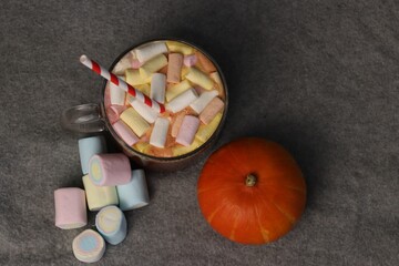 Hot cocoa with marshmallows and striped straw next to pumpkin on gray background