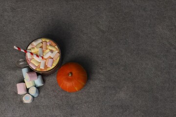 Hot cocoa with marshmallows and striped straw next to pumpkin on gray background