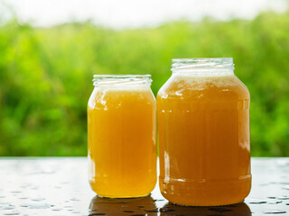 Two glass jars of golden bee honey are on a wet table, green grass field and sky in the background. Fine organic product concept. Agriculture and food industry.