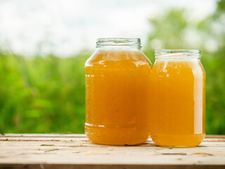 Two glass jars of golden bee honey are on a wet table, green grass field and sky in the background. Fine organic product concept. Agriculture and food industry.