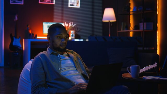 Black guy does remote work from his living room with the laptop, checking online data on screen. Teleworker analyzing and comparing business revenue numbers, focus at night. Camera A.