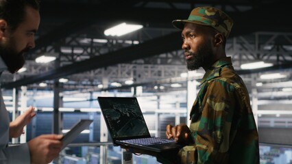 Military technician in data center monitoring warfare drone telemetry on laptop dashboard. IT specialist in army server room using tactical interface on notebook to support aerial operations