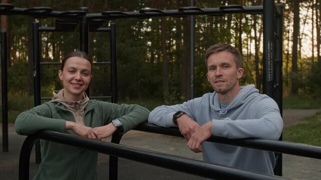 Slow motion portrait shot of two young calisthenics athletes posing for camera, leaning on parallel bars in outdoor gym in forest