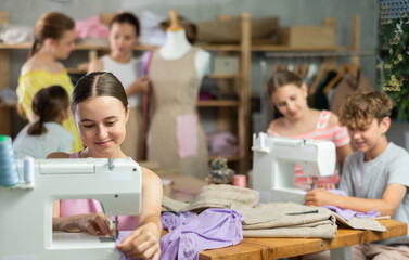Teenage girl works on sewing machine while teacher and students learn how to cut fabric