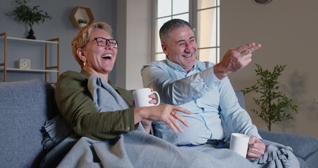 Elderly family couple spending leisure time at home in the evening, watching television and laughing together. The moment highlights their joy and relaxation with TV during a peaceful evening. - Powered by Adobe