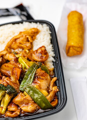 Asian Takeout Meal with Chicken Stir-Fry and Egg Roll on White Background. Black tray filled with chicken stir-fry, vibrant vegetables, and steamed rice, paired with a golden egg roll wrapped in paper