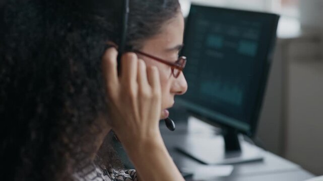 Tilt up of young multiethnic female operator of call center typing on computer keypad, then taking headset, putting it on her head and talking to online client