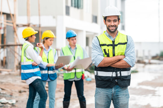 Smiling confident workers, engineers wearing hard hats standing with arms crossed with building team in background at housing construction site