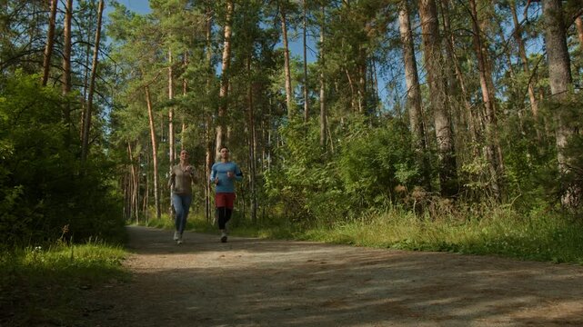 Low angle shot of young man and woman, wearing sport clothes, jogging together down narrow path in forest on sunny day