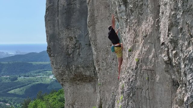 AERIAL: Equipped male rock climber is swiftly climbing a limestone rock face. Surrounding Karst landscape, with distant valley and Adriatic Sea, provides a picturesque backdrop for challenging climb.